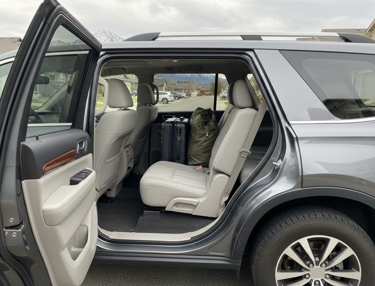 A grey SUV with its rear side door open in a driveway, light grey second-row seats and two bags, a hard-shell suitcase and a green duffel, behind them, with houses and snowy peaks outside